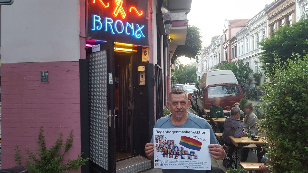 Heinz Hug steht vor der Bronx Bar und hält das Regenbogenmasken Aktionsschild vor sich