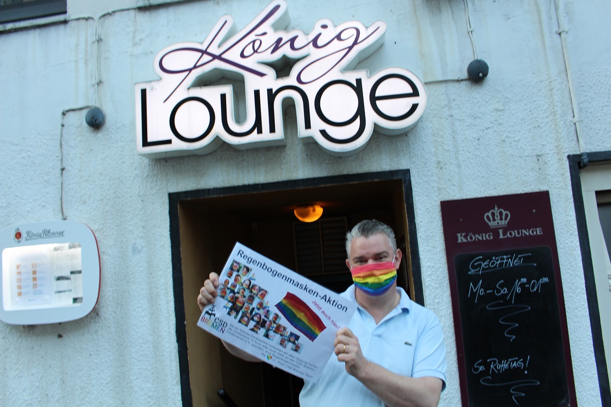 Robert Noetzel steht vor der König Lounge und hält das Regenbogenmasken Aktionsschild hoch
